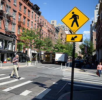 A photograph of a New York City intersection with an enhanced crossing in the form of high-visibility crosswalk markings and a prominent pedestrian warning sign.